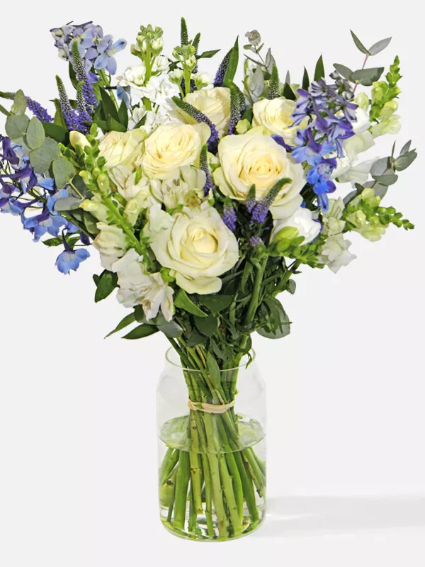 A medium bouquet of white roses, freesia, veronica and blue delphinium in a clear glass vase.